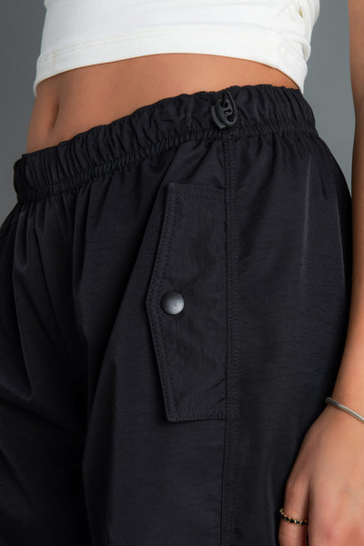 Pantalón parachute de cintura media elástica con doble jareta elástica, fit amplio, plisados frontales y bolsillos frontales decorativos con cartera.