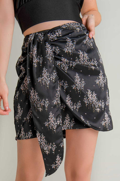Falda corta satinada de estampado floral, corte en a, cintura alta, cruzado frontal con plisado lateral y nudo decorativo. Cierre lateral con cremallera oculta.