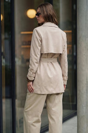 Abrigo trench corto de gamuza, fit recto, cuello solapa, bolsillos delanteros con vivos, detalle de bata frontal y posterior, manga larga con trabillas en puños, cinturón amarrable del mismo tejido y cierre frontal con doble cruce de botones en contraste.