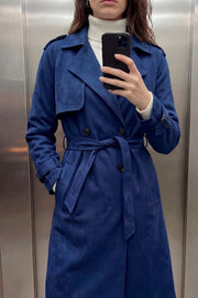 Abrigo trench midi de gamuza, fit recto, con solapa, bolsillos delanteros con vivos, detalle de bata frontal, manga larga con trabillas en puños y hombros, cinturón amarrable del mismo tejido y cierre frontal con doble cruce de botones en contraste.