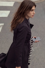 Abrigo trench corto de fit recto, con cuello solapa, bolsillos delanteros con vivos, detalle de bata trasera y manga larga. Trabillas, cinturón amarrable del mismo tejido y cierre frontal con doble cruce de botones en contraste. Detalle de trabilla y botón en hombros, bata trasera, cuello y puños.