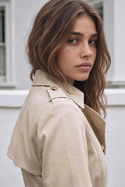 Abrigo trench corto de fit recto, con cuello solapa, bolsillos delanteros con vivos, detalle de bata trasera y manga larga. Trabillas, cinturón amarrable del mismo tejido y cierre frontal con doble cruce de botones en contraste. Detalle de trabilla y botón en hombros, bata trasera, cuello y puños.
