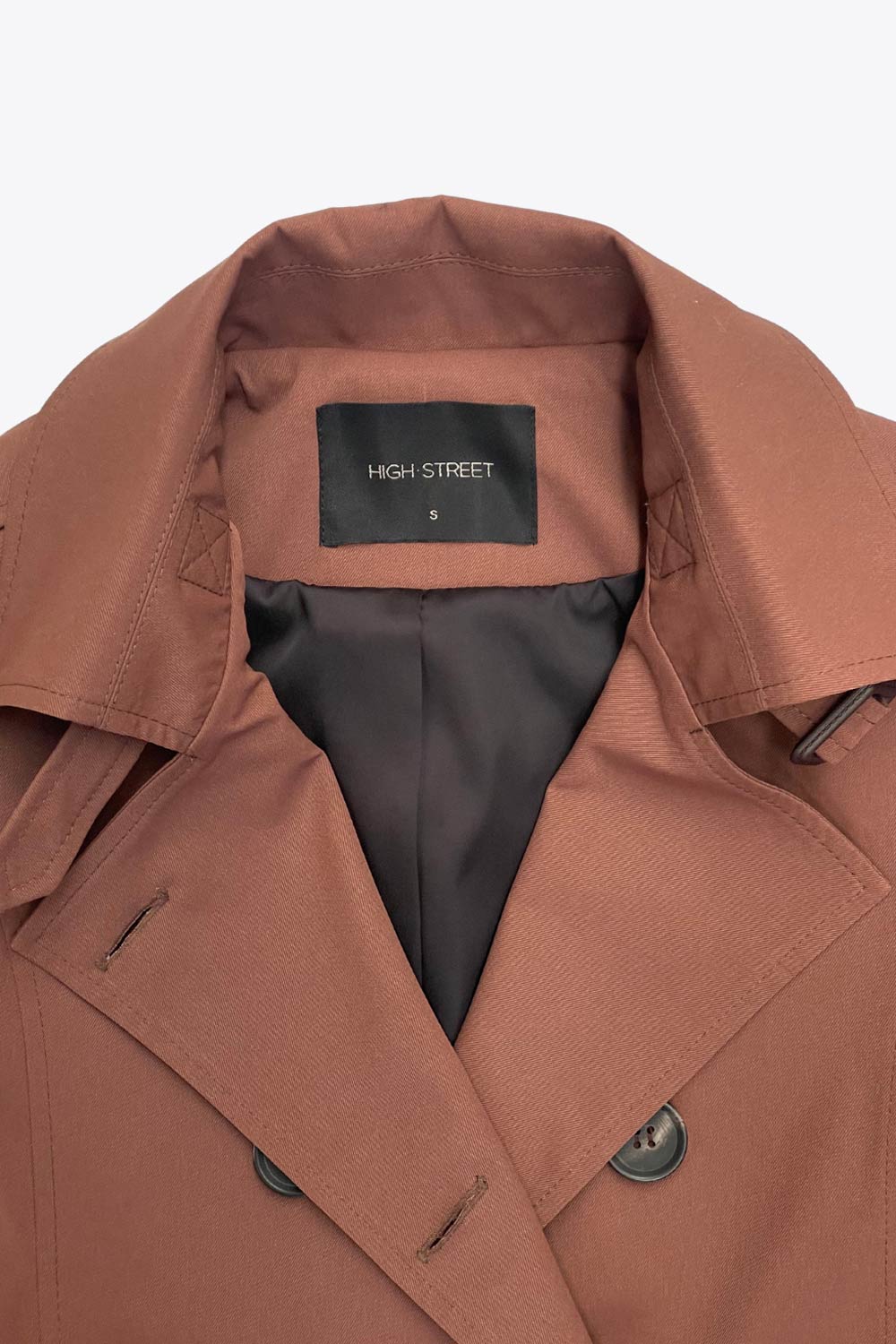 Abrigo trench corto de fit recto, con cuello solapa, bolsillos delanteros con vivos, detalle de bata trasera y manga larga. Trabillas, cinturón amarrable del mismo tejido y cierre frontal con doble cruce de botones en contraste. Detalle de trabilla y botón en hombros, bata trasera, cuello y puños.