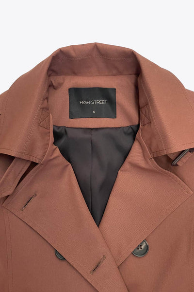 Abrigo trench corto de fit recto, con cuello solapa, bolsillos delanteros con vivos, detalle de bata trasera y manga larga. Trabillas, cinturón amarrable del mismo tejido y cierre frontal con doble cruce de botones en contraste. Detalle de trabilla y botón en hombros, bata trasera, cuello y puños.