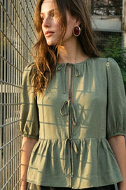Blusa de fit amplio, cuello redondo con plisados, manga corta abullonada con plisados en hombros y rib elástico, peplum con olanes en bajo y cierre frontal con lazos amarrables.