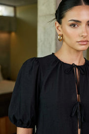 Blusa de fit amplio, cuello redondo con plisados, manga corta abullonada con plisados en hombros y rib elástico, peplum con olanes en bajo y cierre frontal con lazos amarrables.