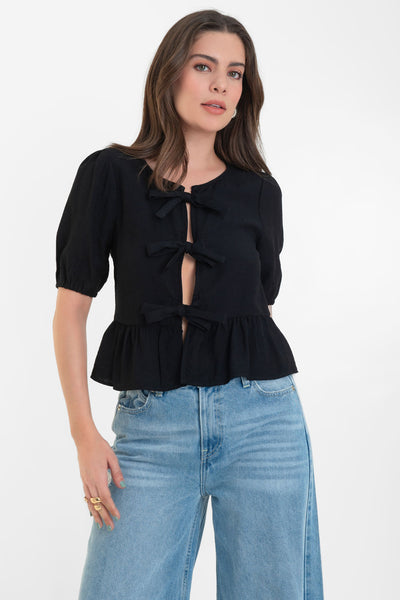 Blusa de manga corta abullonada con rib elástico en bordes, cuello redondo y cierre frontal con lazos amarrables. Detalle de peplum con olanes en bajo.