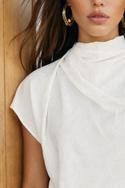 Blusa fluida de manga corta seguida, cuello mock seguido con plisados decorativos en hombro y cuello. Cierre posterior con botones y ojales.