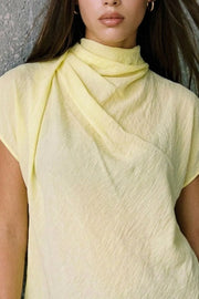 Blusa fluida de manga corta seguida, cuello mock seguido con plisados decorativos en hombro y cuello. Cierre posterior con botones y ojales.