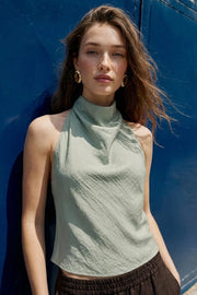 Blusa corta de fit fluido, sin mangas, cuello halter fluido con plisados, escote en espalda y lazo amarrable posterior. Cierre posterior con botón y ojal.