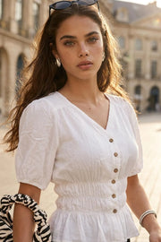 Blusa corta de cuello v, manga corta abullonada, rib elástico con fruncidos en cintura, olanes en bajo y cierre frontal con hilera de botones en contraste.