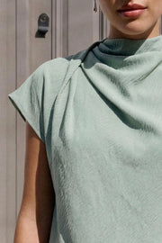 Blusa de cuello alto fluido, plisados en hombros, manga corta seguida y fit recto. Cierre posterior con botón y ojal.