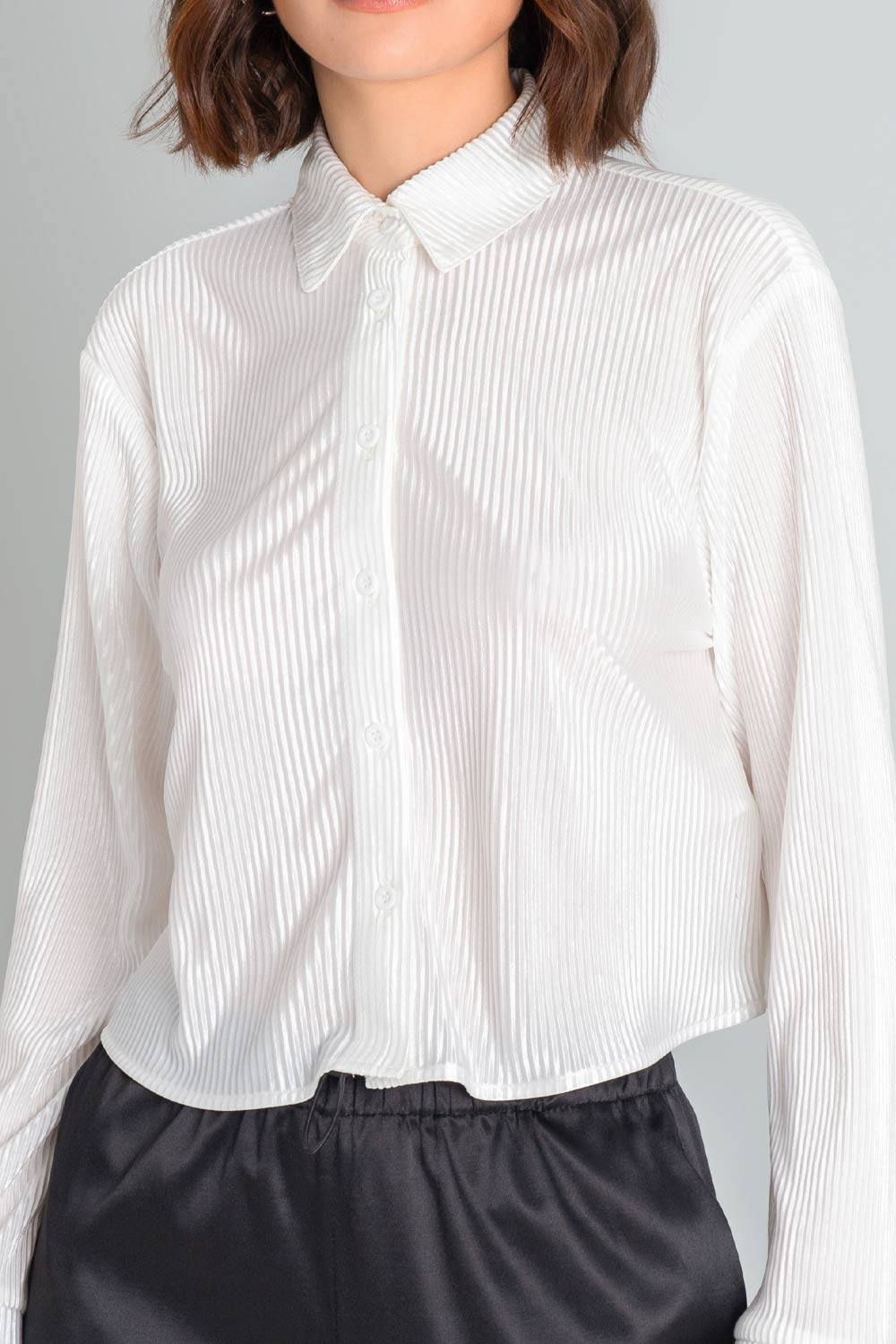Camisa corta satinada de fit recto, manga larga con puño abotonado, cuello camisero y cierre frontal con hilera de botones en contraste. Detalle de plisado en textura de tejido.
