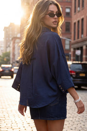 Camisa de fit oversized, manga 3/4 amplia con doblez en puños, cuello camisero, escote v, pinza de amplitud en espalda con trabilla decorativa y bajo curveado. Cierre frontal con hilera de botones en contraste.