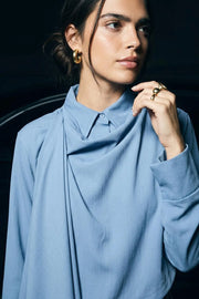 Camisa corta de fit fluido, manga larga amplia con puño abotonado, cuello camisero, detalle decorativo cruzado con plisados y olanes. Cierre frontal con hilera de botones en contraste. Detalle de pinza de amplitud en espalda.