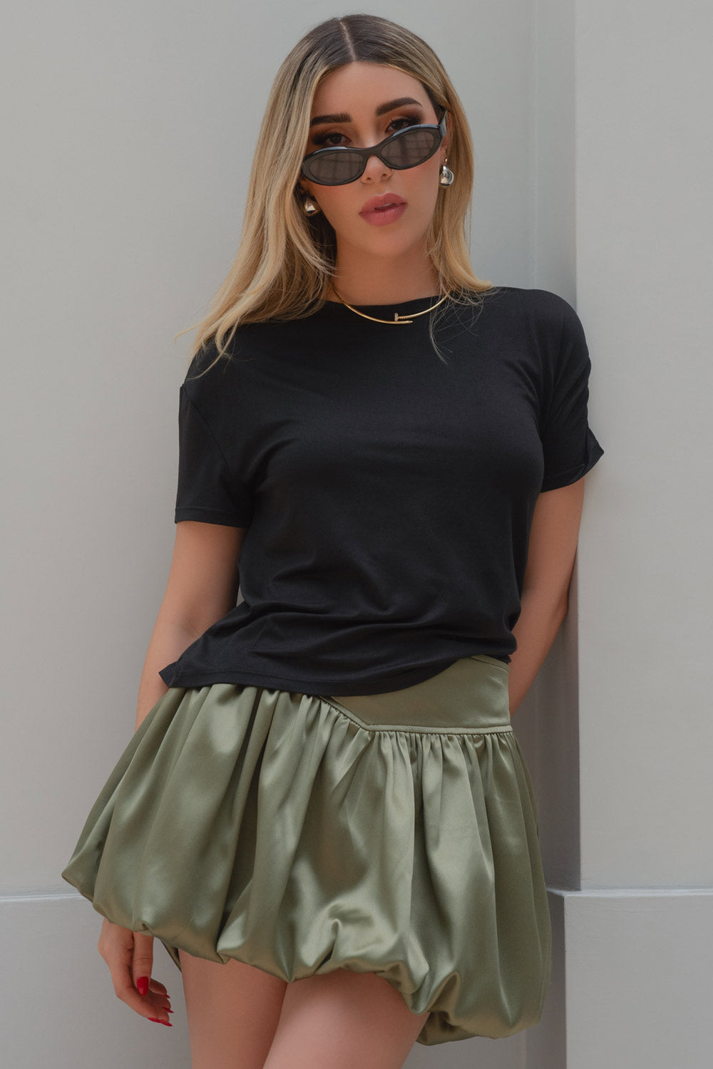 Woman in black t-shirt, olive green skirt, sunglasses, and jewelry, posing against a light-colored wall.