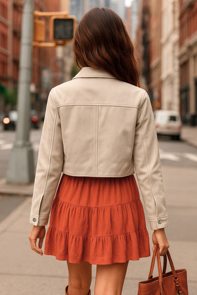 Chamarra corta fluida de gamuza, manga larga con puño abotonado, cuello camisero, bolsillos frontales de parche y cierre frontal con hilera de botones en contraste.