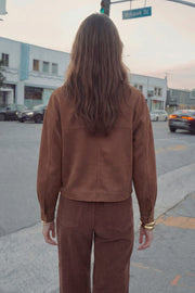 Chamarra corta de gamuza, fit oversized, cuello camisero, manga larga con puño abotonado, corte de bata frontal y posterior, bolsillos frontales de parche y cierre frontal con hilera de botones en contraste.