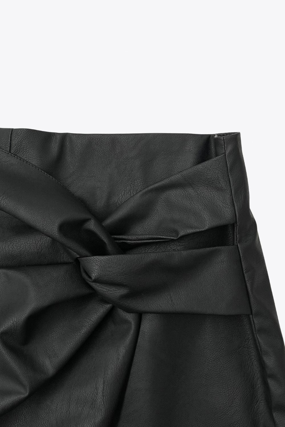 Falda short corto de efecto piel, fit ajustado, cintura alta, cruce frontal asimétrico con nudo y plisados decorativos frontales. Cierre posterior con cremallera oculta.