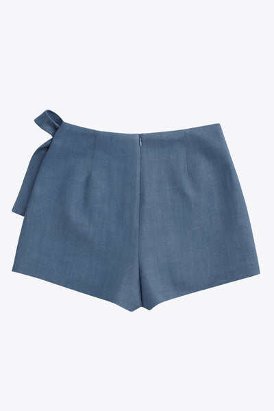 Falda short corto de cintura alta, corte en a, sobrefalda con cruce frontal, lazo lateral amarrable y cierre posterior con cremallera oculta.