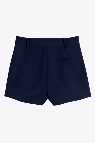 Falda short corto tableado de cintura alta con pretina y trabillas, corte en a, pinzas frontales y vivo decorativo posterior. Cierre frontal con broche y cremallera ocultos.