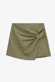 Falda short de fit ajustado, cintura alta, nudo frontal con plisados decorativos y bajo asimétrico. Cierre posterior con cremallera oculta.