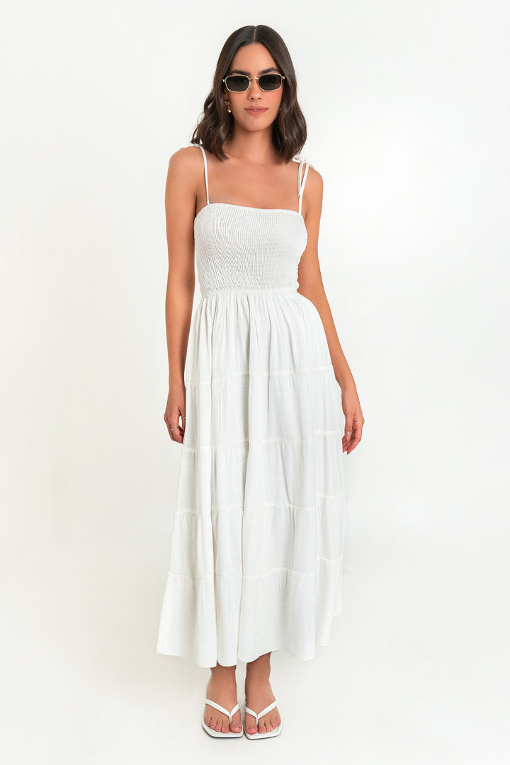 Woman in white sundress and sunglasses standing against a plain background.