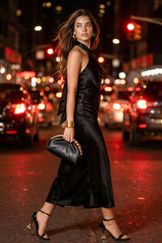 Maxi vestido satinado de fit recto, cuello halter cruzado con lazo amarrable posterior, escote en espalda y cierre posterior con cremallera oculta. Detalle de abertura lateral en bajo.