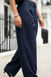 Pantalón de fit wide leg, pinzas frontales, cintura alta con pretina y trabillas, bolsillos delanteros y vivos decorativos traseros. Cierre frontal con broche y cremallera ocultos.