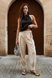 Pantalón de fit balloon, pinzas frontales, cintura alta con pretina, trabillas y cinturón del mismo tejido con ojales, bolsillos delanteros y vivos decorativos posteriores. Detalle de plisados en bajo con botones laterales y trabilla larga en cintura. Cierre frontal con botón y cremallera oculta.