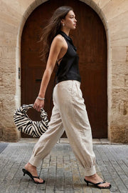 Pantalón de fit balloon, pinzas frontales, cintura alta con pretina, trabillas y cinturón del mismo tejido con ojales, bolsillos delanteros y vivos decorativos posteriores. Detalle de plisados en bajo con botones laterales y trabilla larga en cintura. Cierre frontal con botón y cremallera oculta.