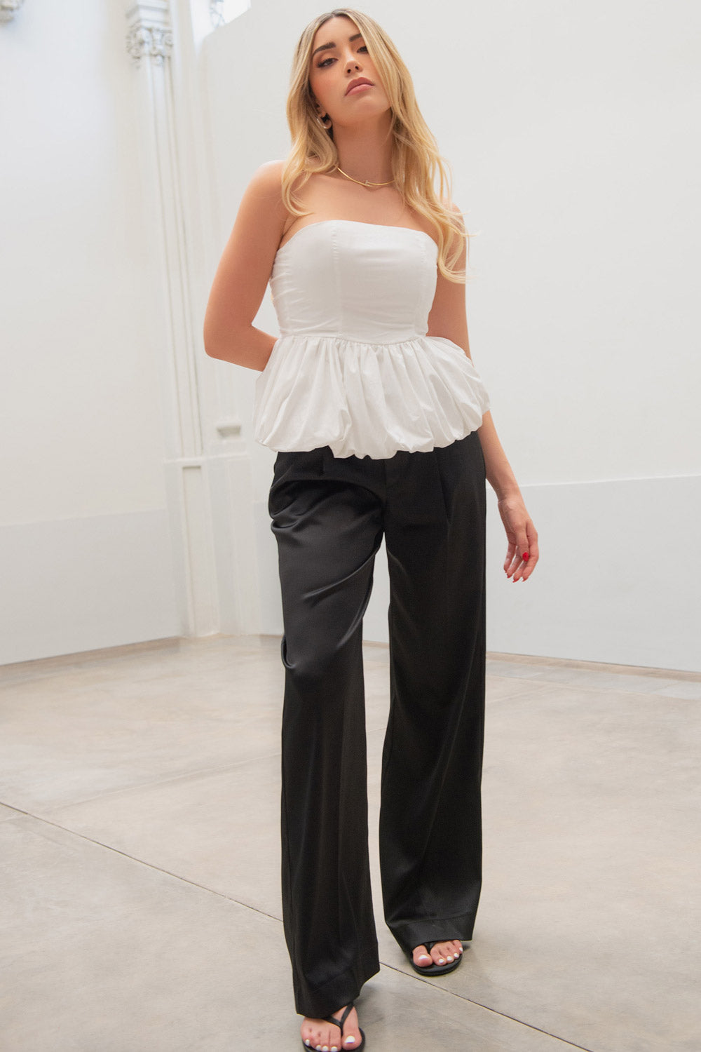 Woman wearing a white strapless top and black wide-leg pants in a minimalist setting.