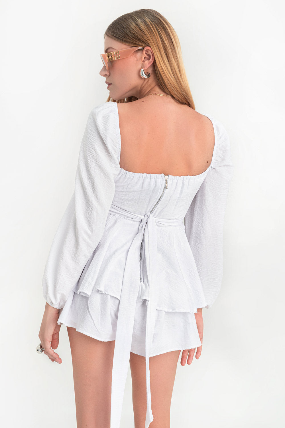 Playsuit de escote v redondeado con detalles plisados, manga larga abullonada con rib elástico en hombros y puños Detalle inferior de capas con olanes, pretina con cruce frontal y lazo amarrable posterior. Cierre con cremallera visible posterior.