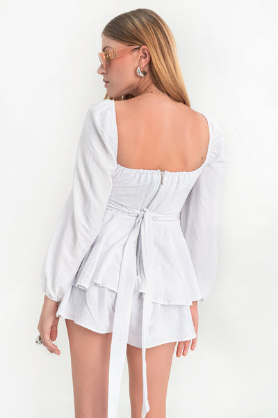 Playsuit de escote v redondeado con detalles plisados, manga larga abullonada con rib elástico en hombros y puños Detalle inferior de capas con olanes, pretina con cruce frontal y lazo amarrable posterior. Cierre con cremallera visible posterior.