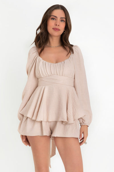 Playsuit de manga larga abullonada con rib elástico en hombros y puños, escote v redondeado con detalles plisados, pretina cruzada con lazo amarrable posterior, capas de olanes en bajo, rib elástico en espalda y cierre posterior con cremallera visible en contraste.