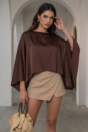 Blusa satinada tipo capa de fit fluido, manga larga amplia, cuello barco y bajo asimétrico.