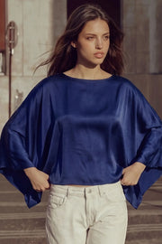 Blusa satinada tipo capa de fit fluido, manga larga amplia, cuello barco, bajo asimétrico y textura en tejido.