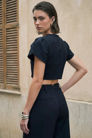 Blusa corta de fit recto, manga corta amplia con abertura, olanes y plisados en hombros, cortes frontales con pinzas, cuello redondo y cierre frontal con hilera de botones en contraste.