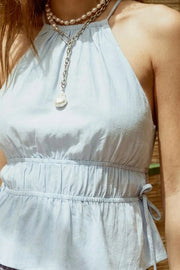 Top corto de lino, cuello halter con plisados, tirantes delgados, detalle de rib con plisados y lazos laterales en costados, peplum con olanes en bajo y cierre posterior con botón y ojal.