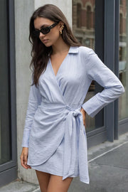 Vestido corto cruzado de estampado rayas, manga larga con puño con abertura decorativa, cuello camisero, escote v, cruce frontal, plisados frontales y nudo amarrable lateral. Detalle de bajo curveado frontal, pinzas de amplitud en espalda y cierre lateral con cremallera oculta.