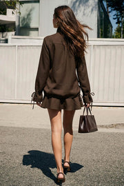 Vestido corto de fit recto, con hombreras, manga larga amplia con jareta y olanes en puños, pinza de amplitud en espalda, bolsillos delanteros con cartera, jareta laterales en bajo con olanes en borde y cierre frontal con doble cruce de botones en contraste. Detalle de cuello alto con trabilla y botón decorativos.